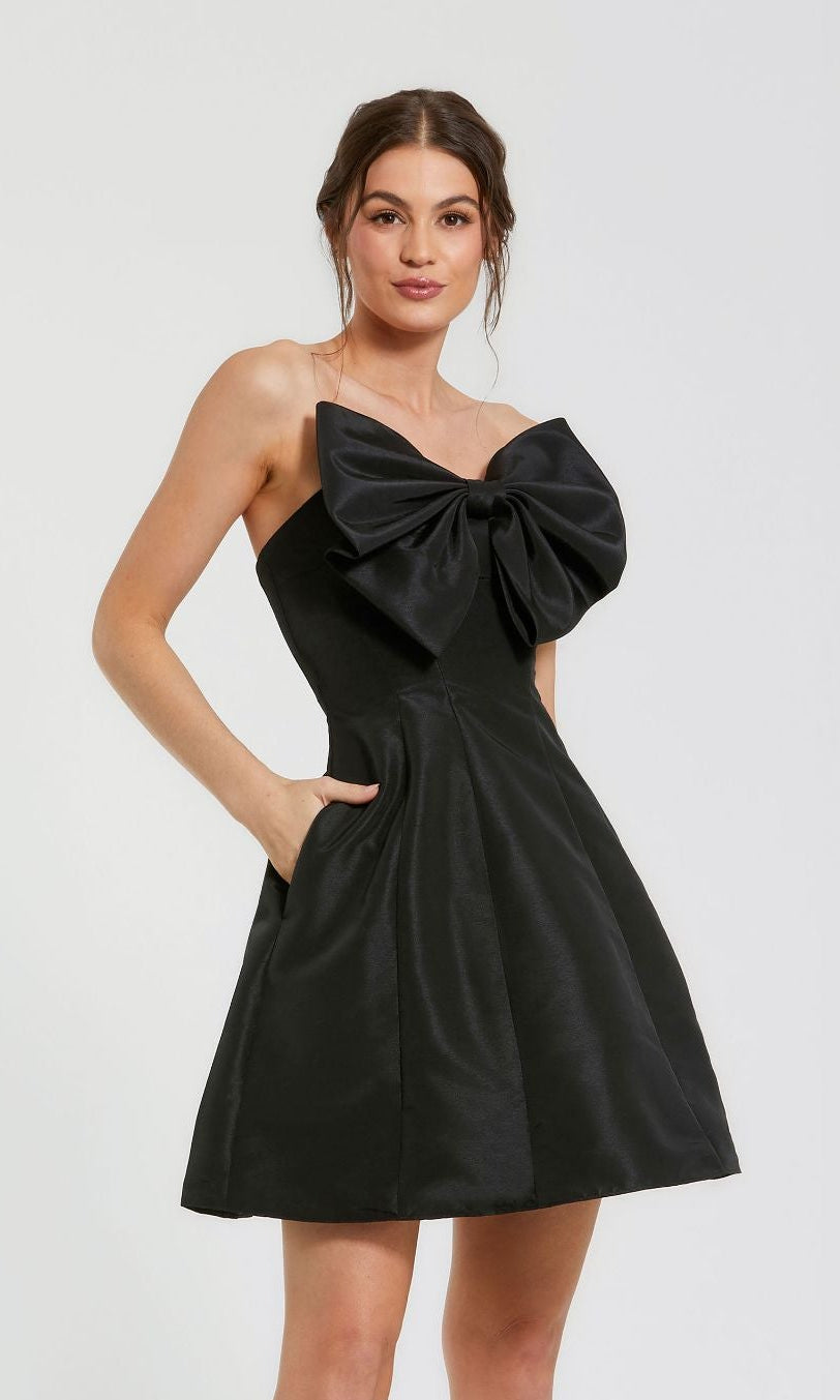 A woman models the Mac Duggal 49210 Strapless Short Bow Party Dress, featuring a large chest bow and pockets, against a plain light background.